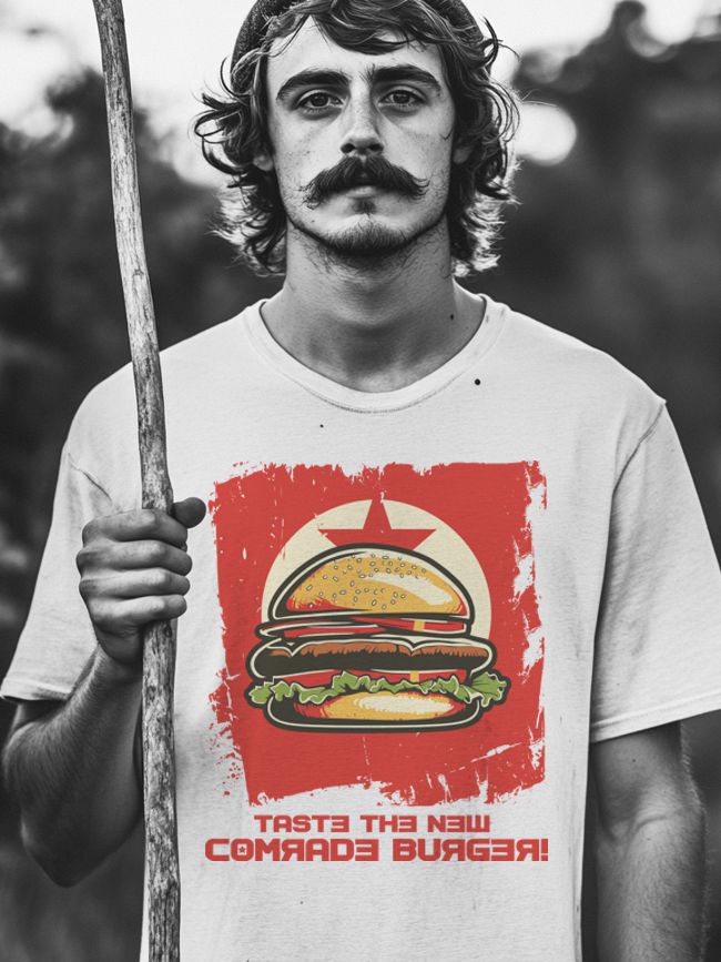 A young man with curly hair and a distinctive mustache holding a wooden stick, dressed in a white t-shirt with a graphic design of a hamburger, and bold text saying 'TASTE THE NEW COMRADE BURGER!' The colors of the t-shirt contrast against a black-and-white filter.